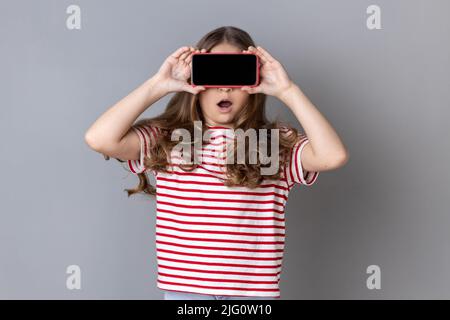 Kleines Mädchen in T-Shirt, die Augen mit Handy bedeckt, posiert mit weit geöffnetem Mund vor Schock Überraschung, unbekanntes Kind versteckt Gesicht mit Handy. Innenaufnahme des Studios isoliert auf grauem Hintergrund. Stockfoto