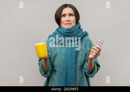 Porträt einer verzweifelten jungen Frau, eingewickelt in einen Schal mit Tee, Serviette, Gefühl der Verzweiflung über Influenza, Tragen einer Jacke im lässigen Stil. Innenaufnahme des Studios isoliert auf grauem Hintergrund. Stockfoto