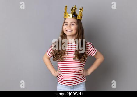 Portrait of satisfied Arrogance kleines Mädchen Prinzessin trägt gestreiftes T-Shirt und goldene Diadem Krone, Blick auf die Kamera hält die Hände auf den Hüften. Innenaufnahme des Studios isoliert auf grauem Hintergrund. Stockfoto