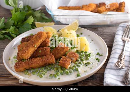 Fischstäbchen mit grünen Erbsen, gekochten Kartoffeln und Bechamelsoße. Serviert auf einem weißen Teller auf einem Holztisch Stockfoto