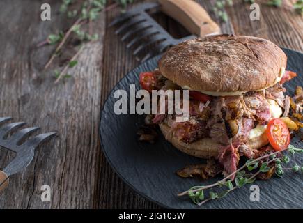 Schweineburger mit Käse, Tomaten und Zwiebeln gezogen. Serviert isoliert auf einem Teller auf rustikalem und hölzernen Tischhintergrund mit Gabeln. Stockfoto