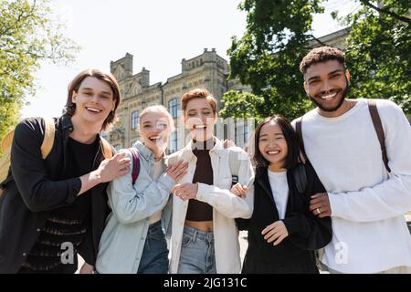 Positive multiethnische Studenten umarmen und betrachten die Kamera im Freien Stockfoto
