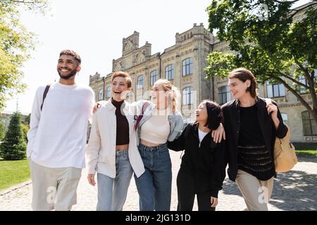 Gruppe lächelnder multikultureller Studenten, die sich umarmen, während sie in der Nähe der Universität im Freien spazieren Stockfoto