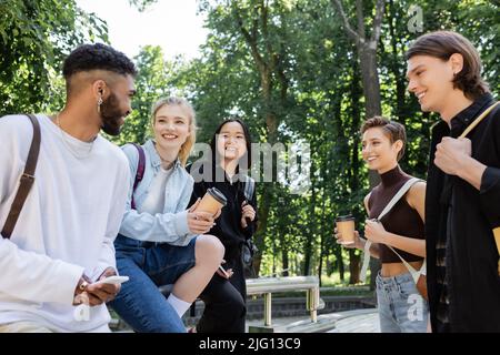 Positive afroamerikanische Studentin mit Smartphone in der Nähe von interrassischen Freunden mit Kaffee im Park Stockfoto