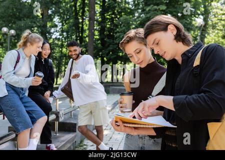 Studenten, die sich ein Notebook mit verschwommenem multiethnischen Freunden und Kaffee und Smartphones im Park angeschaut haben Stockfoto
