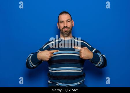 Hispanic-Mann mit gestreiftem Pullover sieht selbstbewusst aus, mit einem Lächeln auf dem Gesicht, das die Finger stolz und glücklich auf sich zeigt Stockfoto
