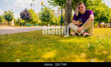 Ein junger Mann mit Dreadlocks ruht im Schatten eines Baumes und nutzt ein Smartphone im Park. Ein gutaussehender Kerl mit Sonnenbrille sitzt auf dem Gras Stockfoto