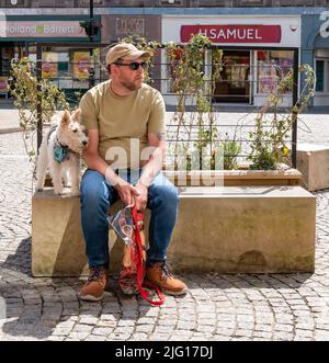 1. Juli 2022. High Street, Elgin, Moray, Schottland. Dies ist ein Mann, der an einem sonnigen Nachmittag mit seinem Hund auf einem Sitz in der Gegend von Elgin sitzt Stockfoto