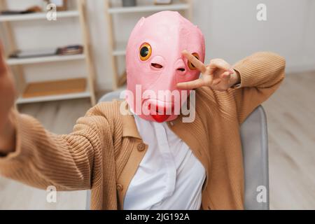 Lustige blonde Dame in rosa Fisch Maske warmen Pullover tun Selfie verwenden zeigen V Zeichen Geste Telefon im Sessel im modernen Zuhause. Pause von der Arbeit, Pause machen Stockfoto