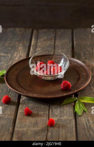 Himbeeren in einer transparenten Untertasse auf einer Keramikplatte auf Holzbrettern. Obst, Essen, Sommer Stockfoto