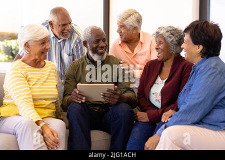 Mehrrassiger älterer Mann mit digitalem Tablet und Blick auf lachende Freunde, während er auf dem Sofa sitzt Stockfoto