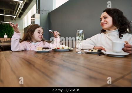 Fröhliche Mutter mit ihrer niedlichen Tochter, die im Flughafenrestaurant schnüffelt, während sie mit dem Flugzeug zusammen in der VIP-Lounge-Cafeteria sitzt und Stockfoto