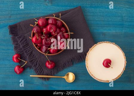 Dunkle und weiße Schokoladenmousse oder Panna Cotta mit Kirschgelee in einem Glas mit Holzschüssel mit reifen Kirschen auf blauem Holztisch. Weitwinkelansicht, n Stockfoto