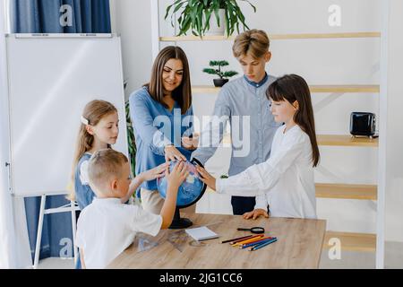 Berühren Sie den Erdball. Eine Gruppe von Kindern unterschiedlichen Alters steht in einem geräumigen Klassenzimmer in der Schule, wobei der Lehrer den Globus berührt. Stockfoto