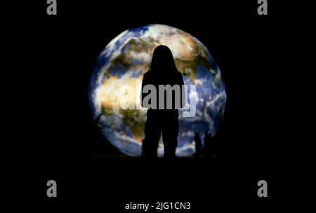 Silhouette einer jungen Frau, die vor einem großen aufblasbaren Modell des Planeten Erde steht, der nachts glüht, Blick von hinten Stockfoto