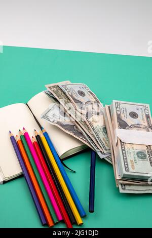 Mehrere Buntstifte auf einem Notizbuch und ein Haufen Dollar, auf einem grünen Tisch mit weißem Hintergrund. Stockfoto