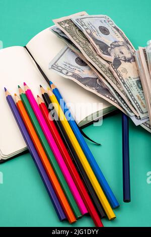 Mehrere Buntstifte auf einem Notizbuch und ein Haufen Dollar, auf einem grünen Tisch mit weißem Hintergrund. Stockfoto