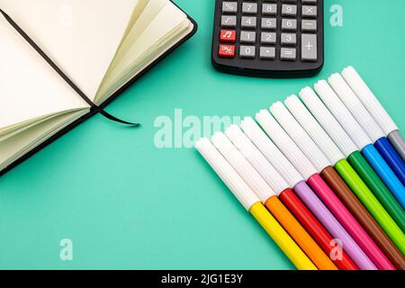 Mehrere Buntstifte auf einem Notizbuch und ein schwarzer Rechner auf einem grünen Tisch. Zurück zur Schule. Stockfoto