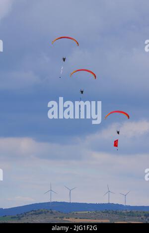 Drei Fallschirmspringer am bewölkten Himmel ziehen Flagge, Banner und Poster von Atatürk für eine Airshow in der Türkei und Windturbinen auf Hügeln im Hintergrund Stockfoto