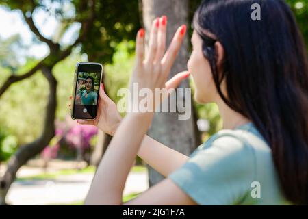 Glückliche Brünette Frau trägt türkisfarbenes T-Shirt im Stadtpark, im Freien ein Selbstporträt mit Smartphone. Sie schaut auf den Bildschirm und macht Selfie. Stockfoto