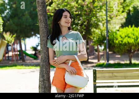 Glückliche Frau mit T-Shirt, die im Stadtpark steht, sich im Freien gegen einen Baum lehnt und für die Kamera posiert. Eine Handtasche halten und mit großem SM wegschauen Stockfoto