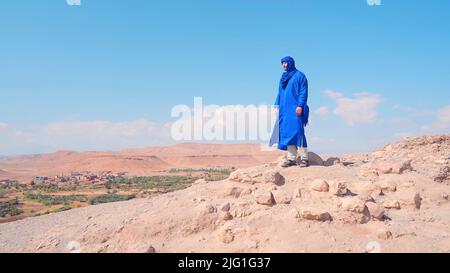 Staubige Wüstenstraße mit niedrig wachsenden Büschen und Pflanzen auf blauem Hintergrund. Aktion. Ein Mann in blauer männlicher arabischer Kleidung, der auf einem Hügel steht Stockfoto