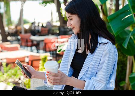 Seitenansicht einer niedlichen Millennial-Frau, die im Stadtpark ein Hemd trägt, im Freien einen Kaffeebecher hält und ein Smartphone verwendet. Blättern o Stockfoto