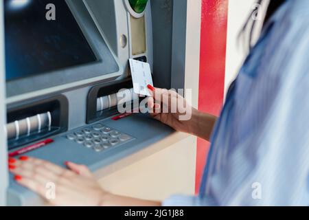 Schöne Brünette Frau trägt gestreiftes Hemd auf Stadtpark stehen, im Freien vor dem Geldautomaten mit Kredit- oder Debitkarte stehen. Stockfoto