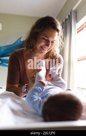 Lächelnde kaukasische Mutter mit mittlerem Erwachsenen, die ihr neugeborenes Baby auf dem Wickeltisch zu Hause ankleidet Stockfoto