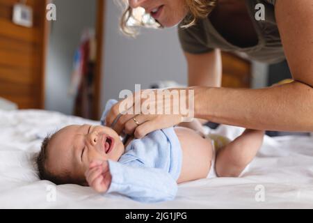 Kaukasische Mutter mit mittlerem Erwachsenen, die weinendes Neugeborenes zu Hause auf dem Bett kleidet, Platz kopieren Stockfoto