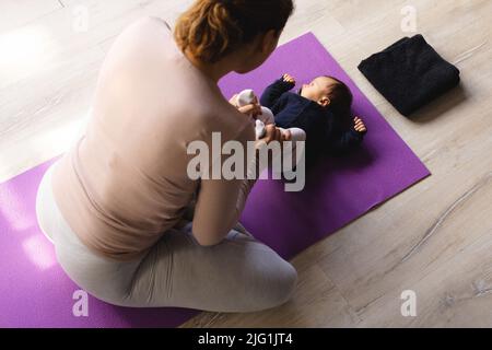 Hochwinkelansicht der kaukasischen Mutter mit mittlerem Erwachsenen, die zu Hause ein niedliches neugeborenes Baby auf einer Yogamatte hält Stockfoto