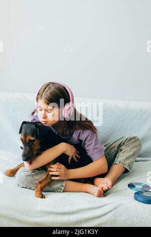 Fröhliches Teenager-Mädchen im lila T-Shirt in rosa Kopfhörern hört Musik und hat Spaß beim Spielen mit Hund Stockfoto