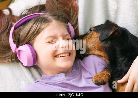 Fröhliches Teenager-Mädchen im lila T-Shirt in rosa Kopfhörern hört Musik und hat Spaß beim Spielen mit Hund Stockfoto