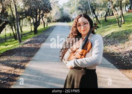 Lächelnde Brünette Frau mit einer Brille, die draußen in einer Parkstraße eine Geige hält und umarmt. Stockfoto