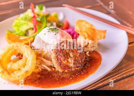 Hawaiianische Loco Moko Reisplatte mit Gemüse, einem Hamburger, einem Spiegelei und gebratenen Zwiebelringen auf einem Teller mit Essstäbchen in einem japanischen Restaurant von Stockfoto
