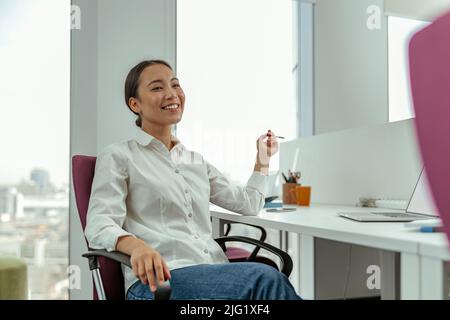 Lächelnde asiatische Geschäftsfrau sitzt im Büro an ihrem Arbeitsplatz und schaut weg Stockfoto