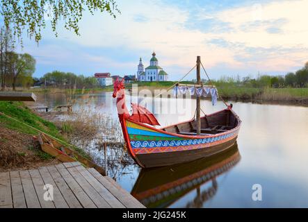 Tourist 'drakkar' auf dem Fluss. Ein Boot am Ufer des Flusses Kamenka, Elijaskirche am Horizont. Susdal, Russland, 2022 Stockfoto