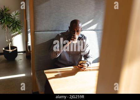 afroamerikanischer reifer Geschäftsmann, der während der Büropause ein Smartphone verwendet, während er Kaffee getrunken hat Stockfoto