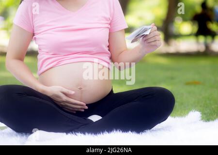 Nahaufnahme Glück junge asiatische Frau schwanger sitzen berühren Bauch und suchen Foto mit positiven im Park, Mutter und Gesundheit der Schwangerschaft, glücklich Stockfoto