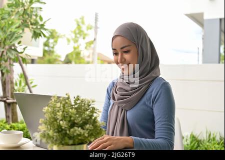 Schöne tausendjährige asiatische muslimische Frau mit Hijab sitzt in schönen Garten zu Hause und mit tragbaren Laptop-Computer. Stockfoto