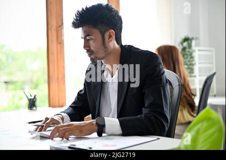 Intelligenter, professioneller junger, erwachsener asiatischer Geschäftsmann oder Büroangestellter, der an seinem Finanzprojekt im Büro arbeitet. Stockfoto