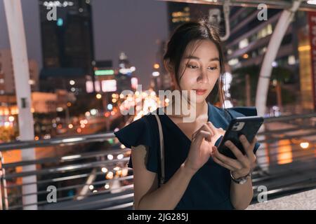 Schöne junge asiatische Frau, die über eine Fußgängerbrücke steht und Smartphone überprüft und Apps durchsucht, während sie E-Mails abcheckt Stockfoto