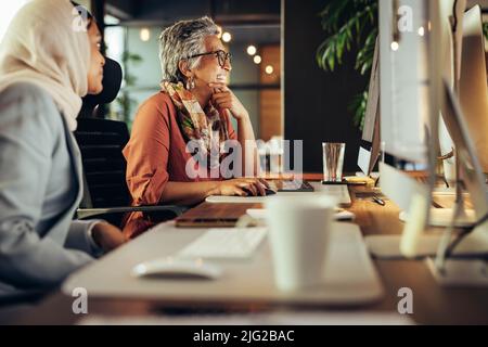 Zwei Geschäftsfrauen lächeln fröhlich, während sie in einem Büro auf einen Computerbildschirm blicken. Diverse Geschäftsfrauen arbeiten als Team in einem kreativen Co-Working Stockfoto