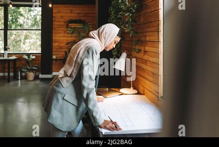 Nachdenklicher muslimischer Designer, der in einem modernen Büro mit Zeichnungen arbeitet. Kreativer junger Architekt, der ein neues innovatives Projekt plant. Junge Geschäftsfrau w Stockfoto