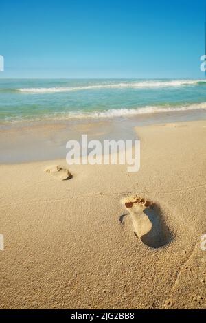 Schritte an einem Sandstrand am Meer mit einer weißen schäumenden Welle aus dem blauen Meer. Weiche Wellen von klarem Wasser. Sandstrand mit menschlichen Fußabdrücken. Angenehm Stockfoto