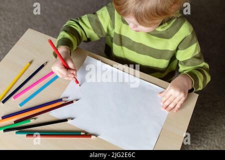 Kleine Kinderzeichnung mit Buntstiften auf weißem Papier, die am Tisch sitzt. Draufsicht. Stockfoto