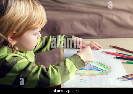 Kleines Kind zeichnet mit Buntstiften auf weißem Papier und sitzt zu Hause am Tisch Stockfoto