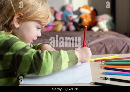 Kleines Kind Zeichnung mit Buntstiften auf weißem Papier sitzen am Tisch zu Hause mit Spielzeug auf dem Bett im Hintergrund Stockfoto