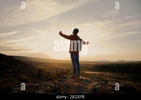 Junger Mann mit Rucksack, der mit ausgestreckten Händen auf dem Berg steht Stockfoto