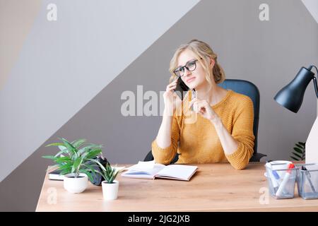 Glückliche Geschäftsfrau, die am Mobiltelefon anruft und Notizen auf einem Schreibtisch im Büro macht, während sie auf einem gelben Teppich sitzt und eine Brille trägt. Kaukasische Frau bei der Arbeit Stockfoto